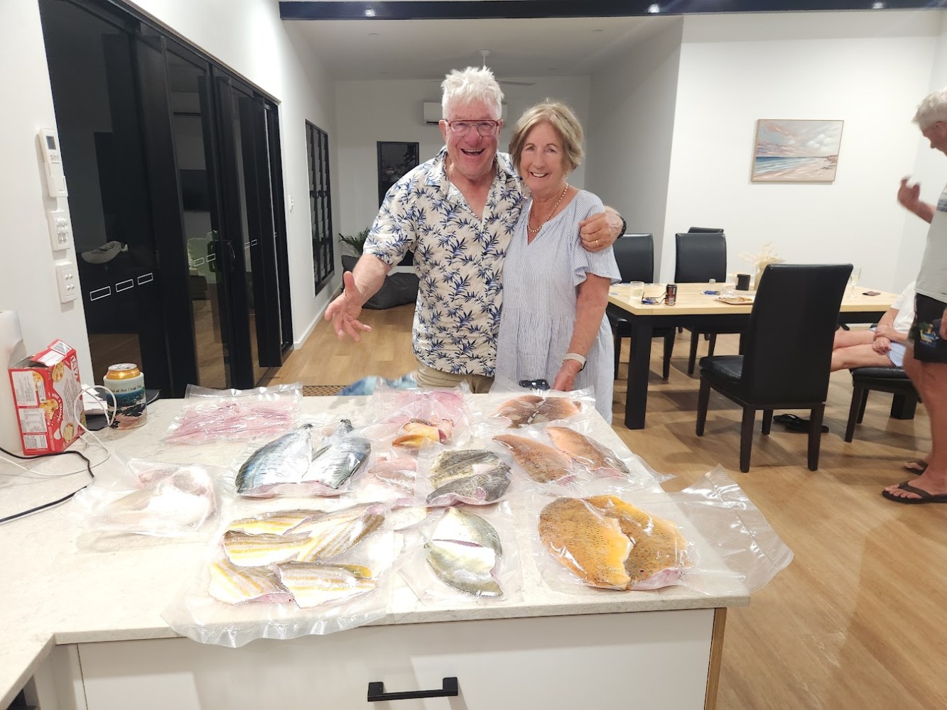 Smiling couple poses with vacuum-sealed fish fillets spread on a kitchen counter.
