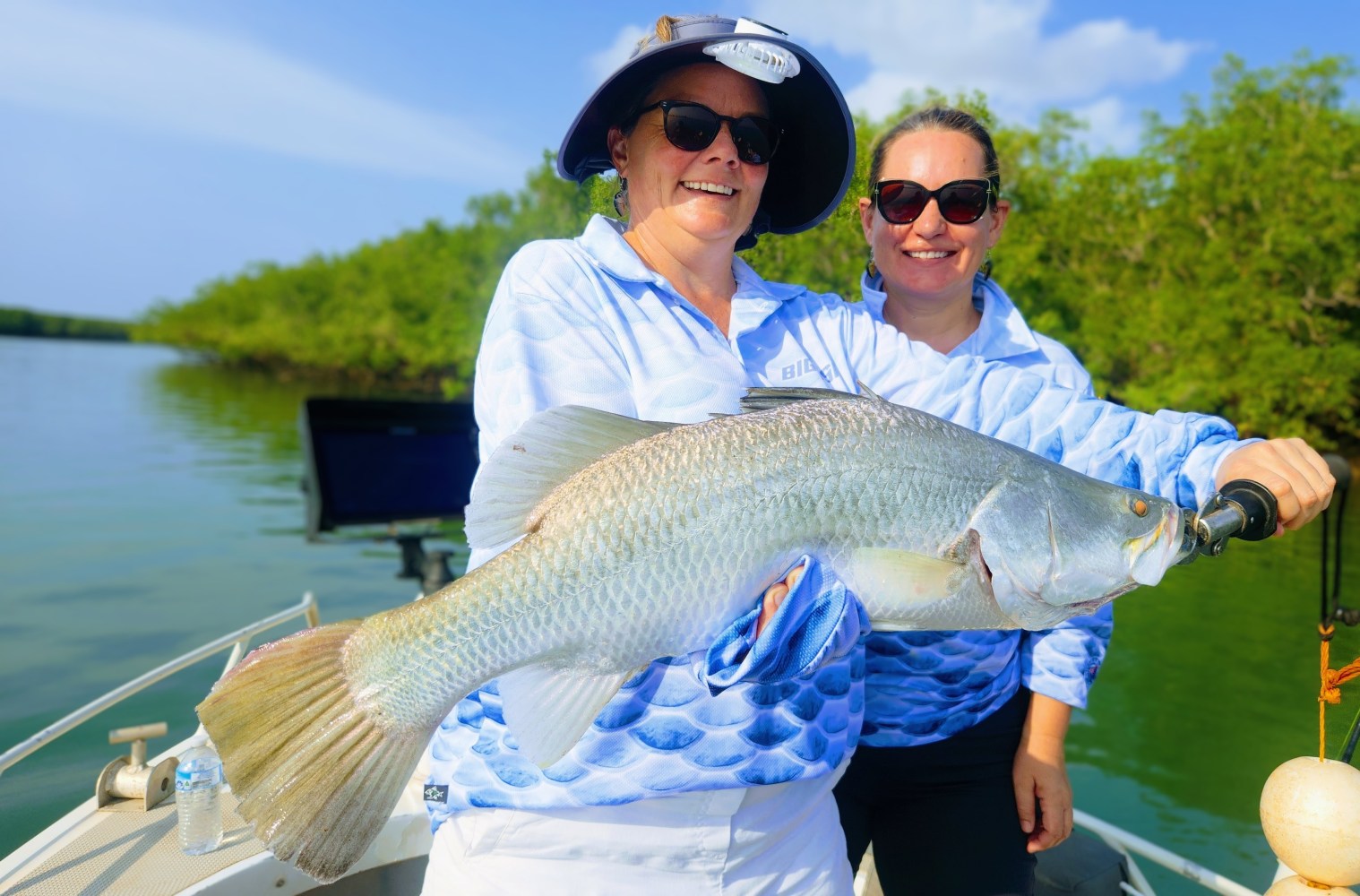Two people in sunglasses holding a large fish on a boat with trees in the background.