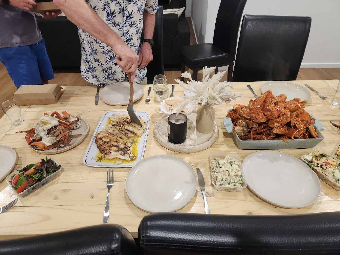 Seafood meal on table with plates of crabs, fish, and side dishes. Two people nearby, one serving food.