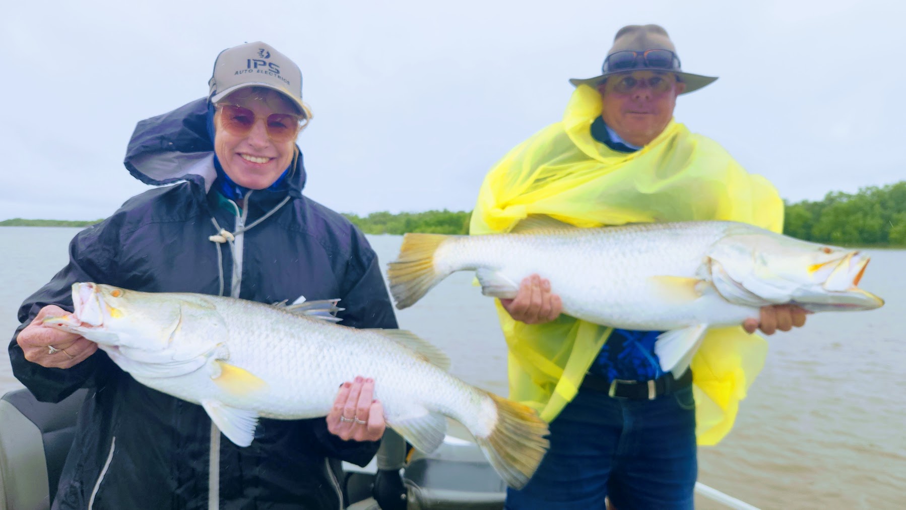 Two people on a boat holding large fish, wearing rain jackets and hats.