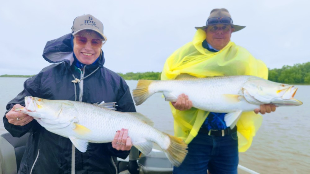 Two people on a boat holding large fish, wearing rain jackets and hats.