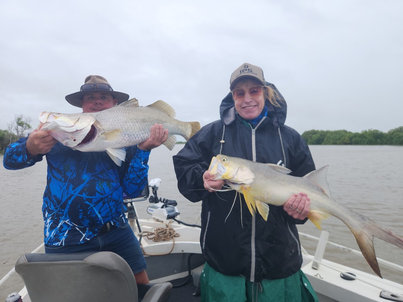 Two people on a boat holding large fish against a cloudy sky.