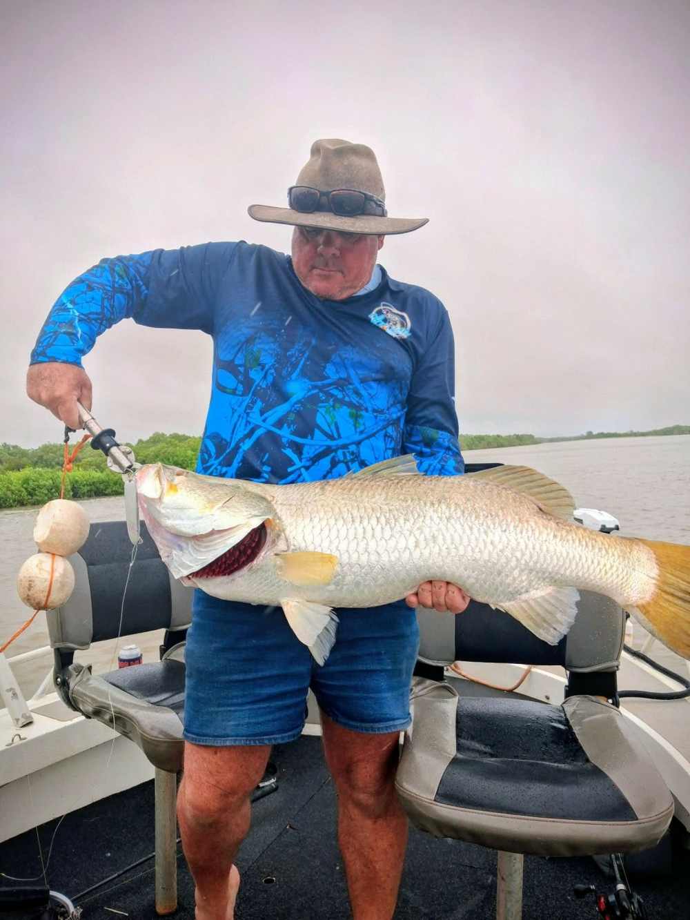 Man on a boat wearing a hat holds a large fish with a fishing grip.
