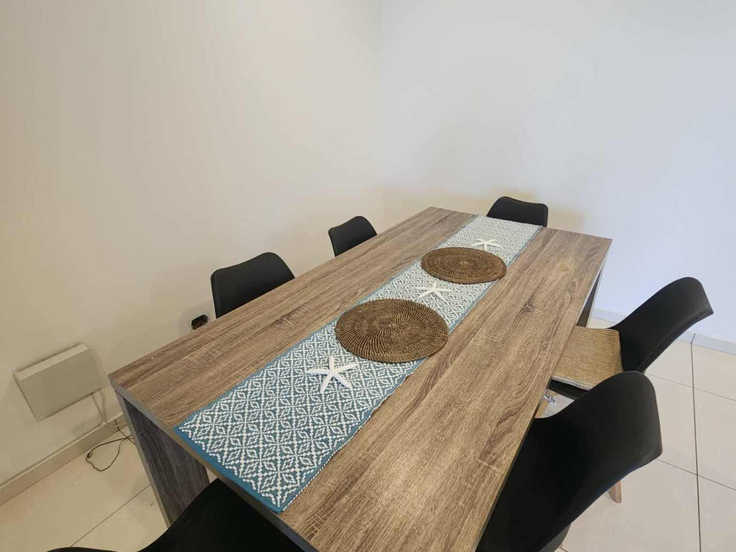 Wooden table with six black chairs, placemats, and a blue runner in a minimal dining room.