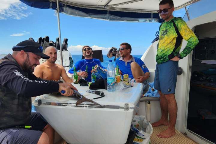 Five men on a boat, one cleaning a fish, others smiling around a table.