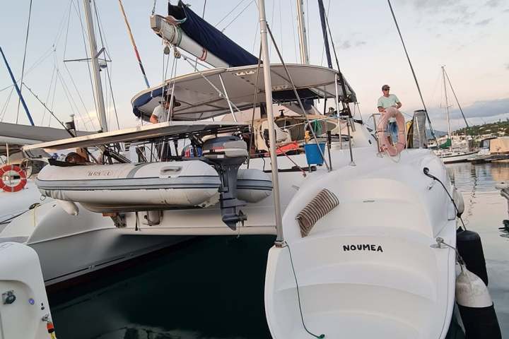 Docked catamaran with sails down, small motorboat, and people onboard.