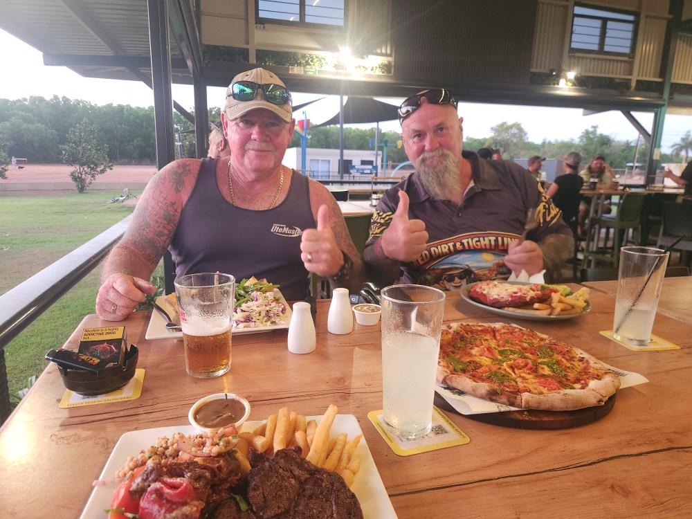 Two men seated at a table with steak and pizza, drinks and condiments visible.