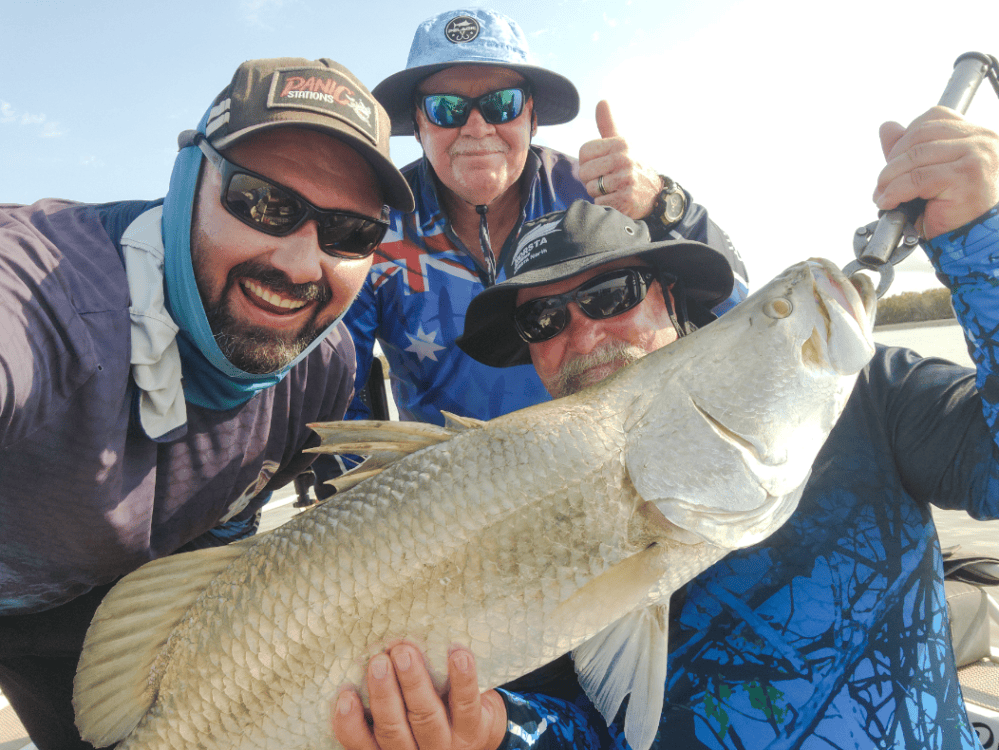 Three men posing with a large fish on a boat, one holding the fish.